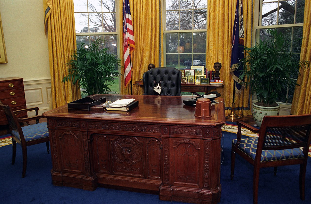 1280px-Photograph_of_Socks_the_Cat_Sitting_Behind_the_President's_Desk_in_the_Oval_Office-_01-07-1994_(6461515323)
